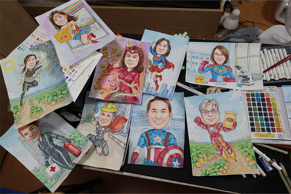 ภาพรวมงานวาดการ์ตูนล้อเลียนแนวซูเปอร์ฮีโร่และอาชีพต่าง ๆ หลายใบวางบนโต๊ะ พร้อมอุปกรณ์วาดภาพ สีน้ำ พู่กัน และมาร์กเกอร์ เห็นบรรยากาศโต๊ะทำงานของช่างเหิน
