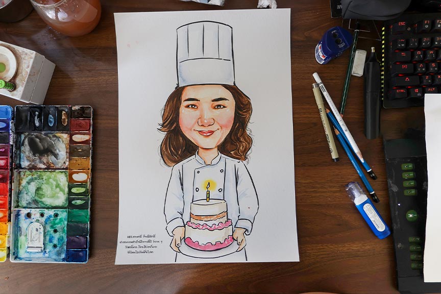 วาดรูปเป็นของขวัญ เหมาะกับโอกาสไหนบ้าง? ภาพวาดการ์ตูนเชฟถือเค้กวันเกิดบนกระดาษ พร้อมสีน้ำและอุปกรณ์วาดรูปบนโต๊ะ